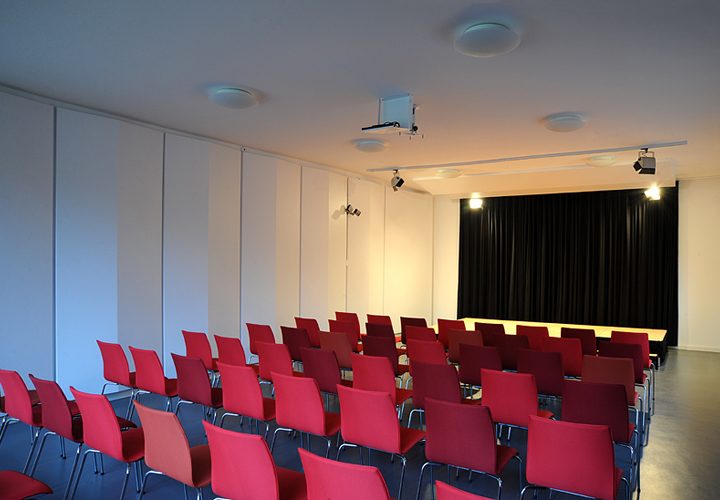 Saal mit roter Bestuhlung und schwarzem Bühnenvorhang